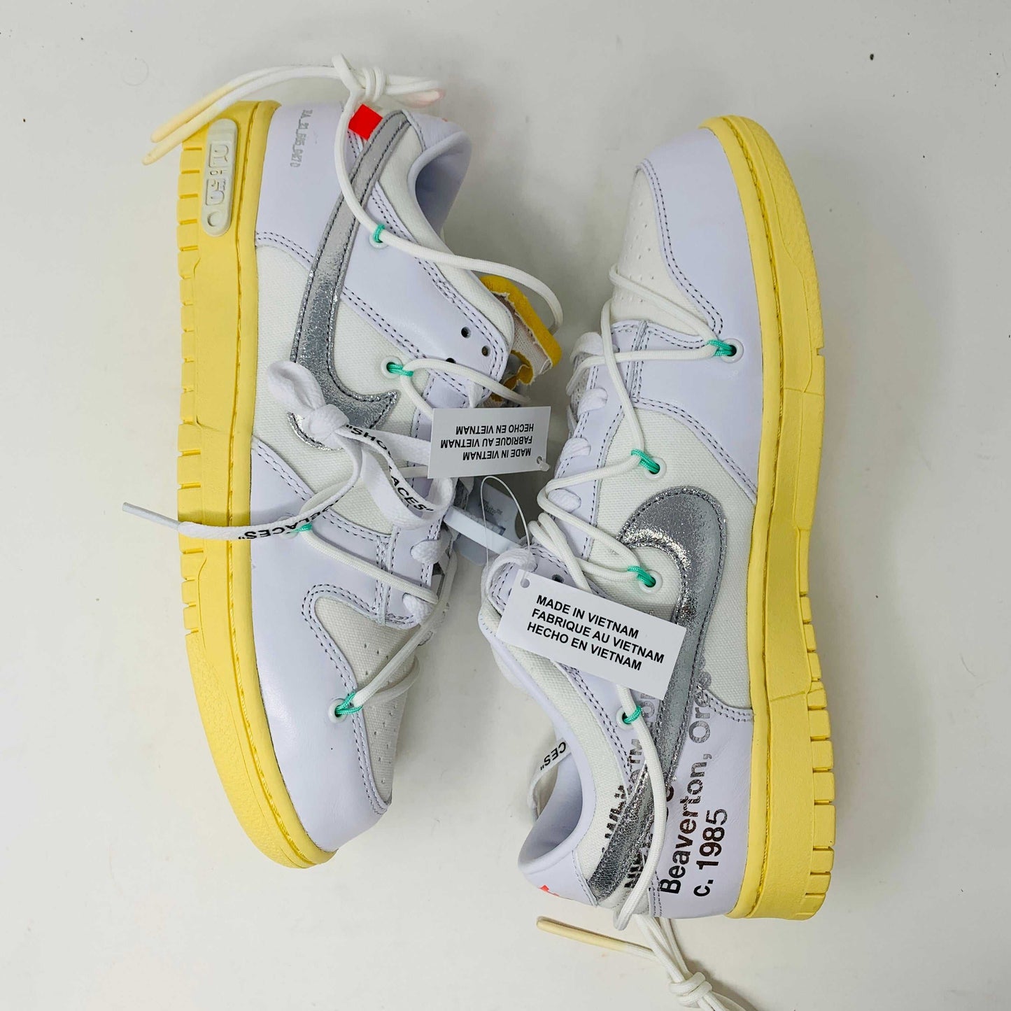 The Nike Dunk Low Off White Lot 1 sneaker, white with a metallic silver swoosh and vibrant accents, rests on a brown shoebox. Its unique lace design and 01 of 50 detailing highlight its 2021 release against a plain white backdrop.