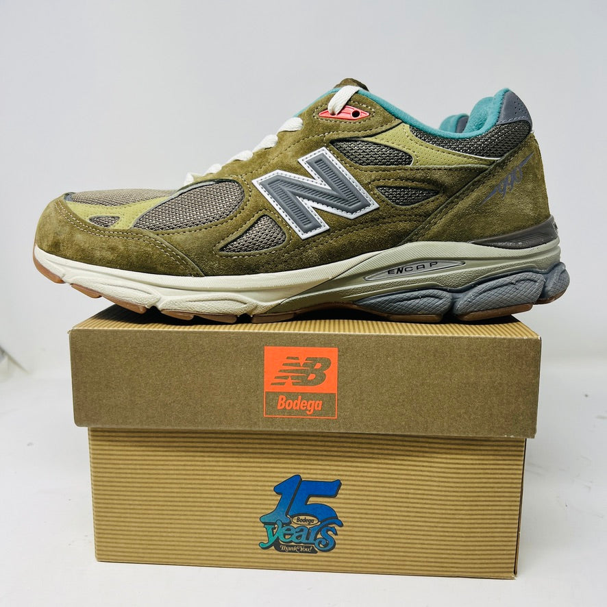 A New Balance 990v3 MiUSA Bodega Here To Stay sneaker in green and gray with white laces, detailed stitching, and side branding sits on a brown box featuring New Balance, Bodega, and 15 Years logos. This 2021 edition is a stylish wardrobe accessory.