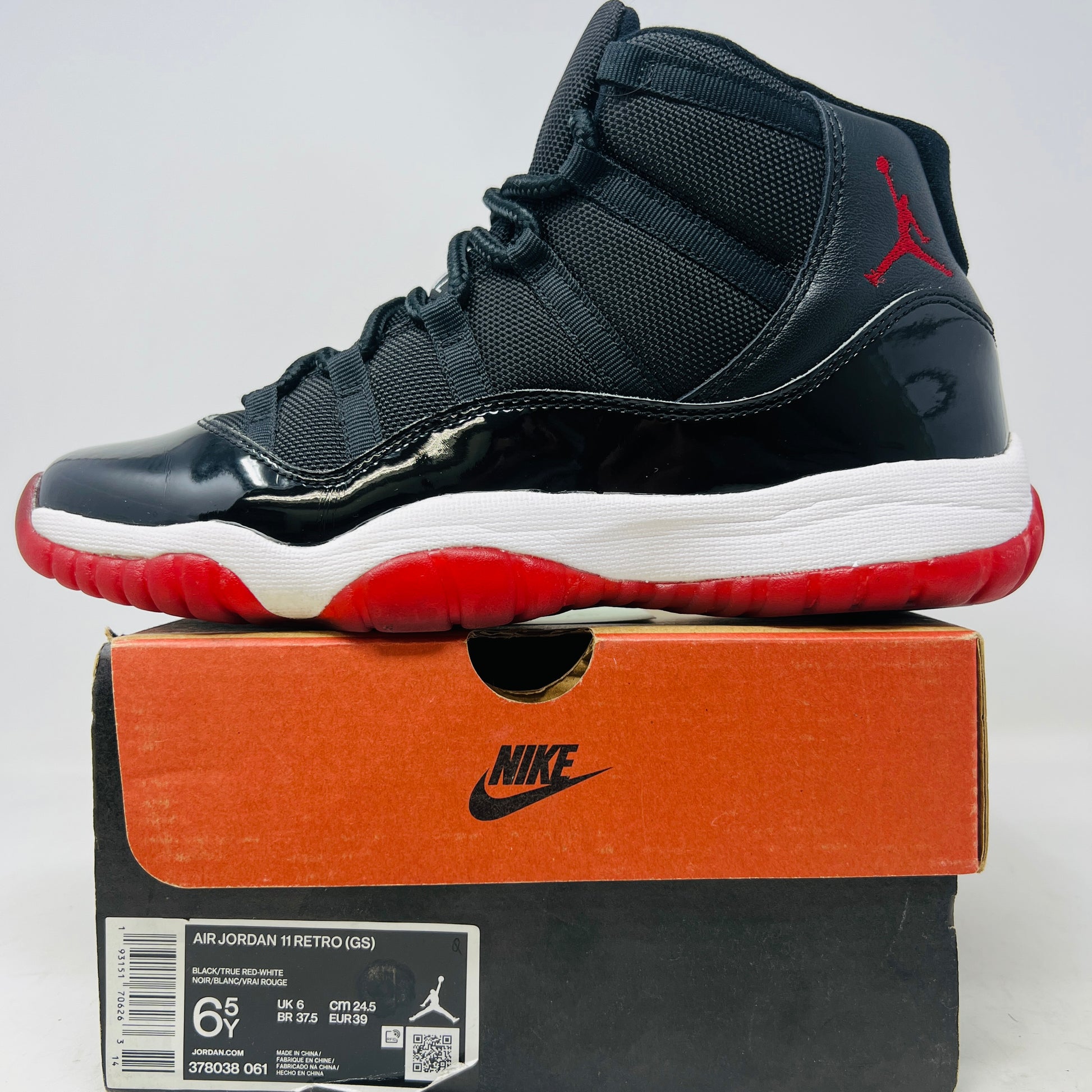 A pristine black and red Jordan 11 Retro Playoffs Bred (2019) (GS) sneaker is on an orange Nike box, featuring a patent leather upper, white midsole, red outsole, and the Jumpman logo near the heel. The label indicates year and size details.