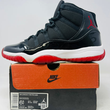A pristine black and red Jordan 11 Retro Playoffs Bred (2019) (GS) sneaker is on an orange Nike box, featuring a patent leather upper, white midsole, red outsole, and the Jumpman logo near the heel. The label indicates year and size details.