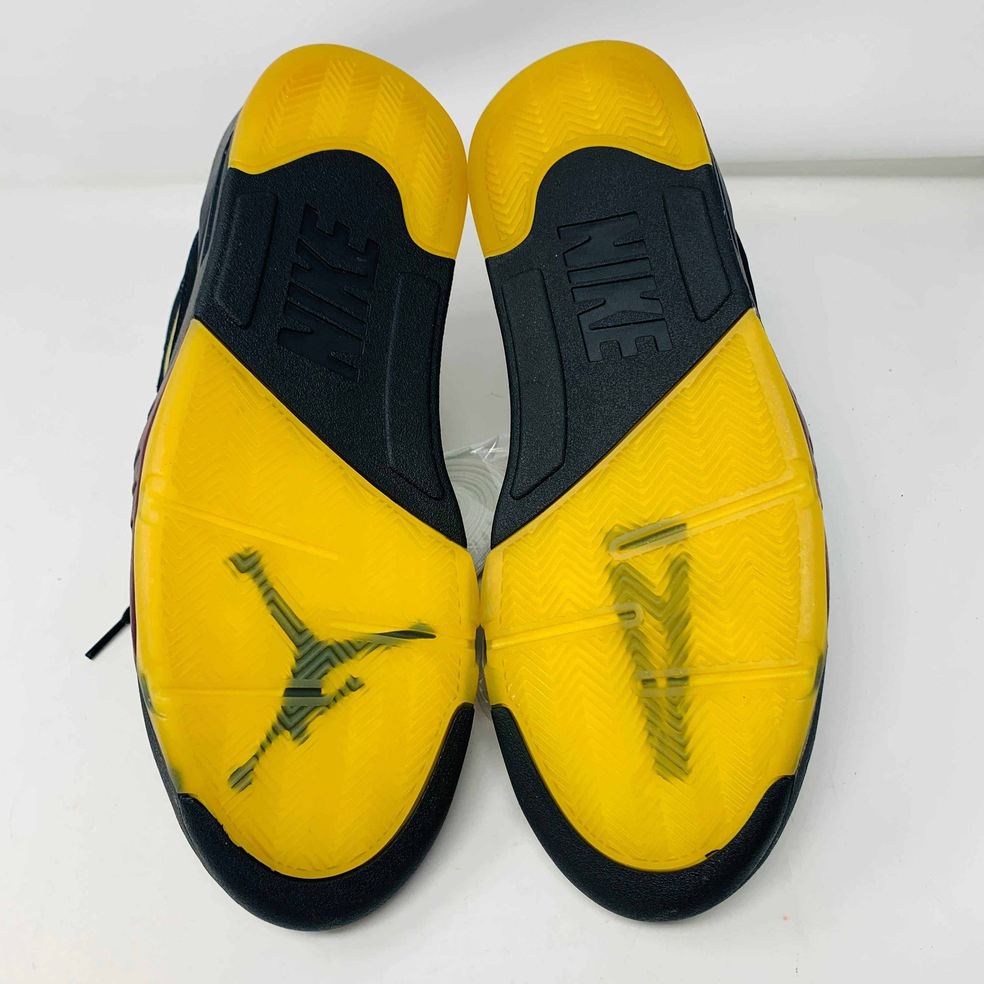 The image displays the soles of Jordan 5 Retro A Ma Maniére Dusk sneakers featuring a black and yellow design. The word NIKE is engraved on the heels, with a black Jumpman logo visible on the translucent yellow section.