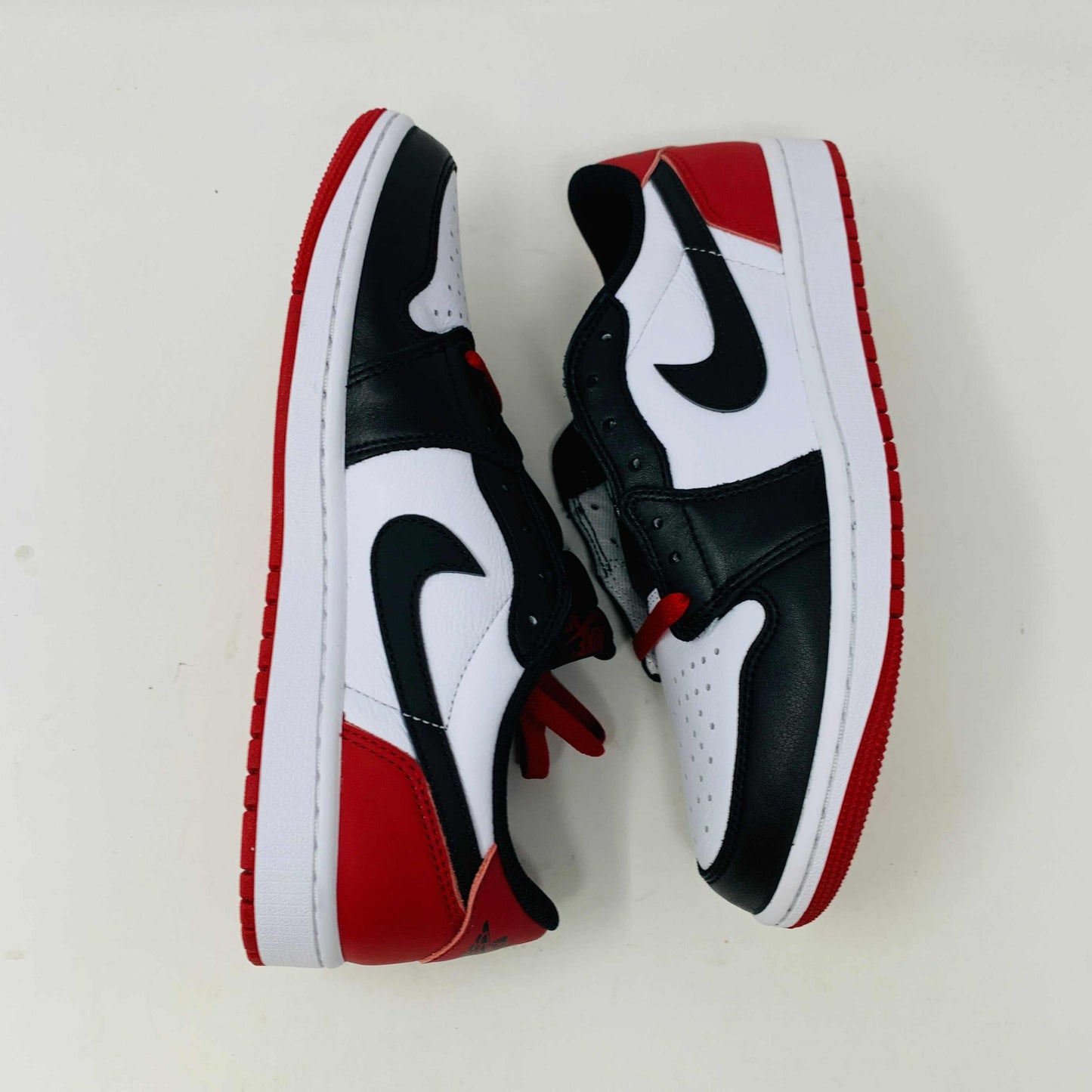 Discover the Jordan 1 Retro Low OG Black Toe (2023) sneakers featuring genuine black and white leather panels, red laces, and soles. These low-tops from Jordan come with extra laces and are displayed on a clean white surface against a pristine background.