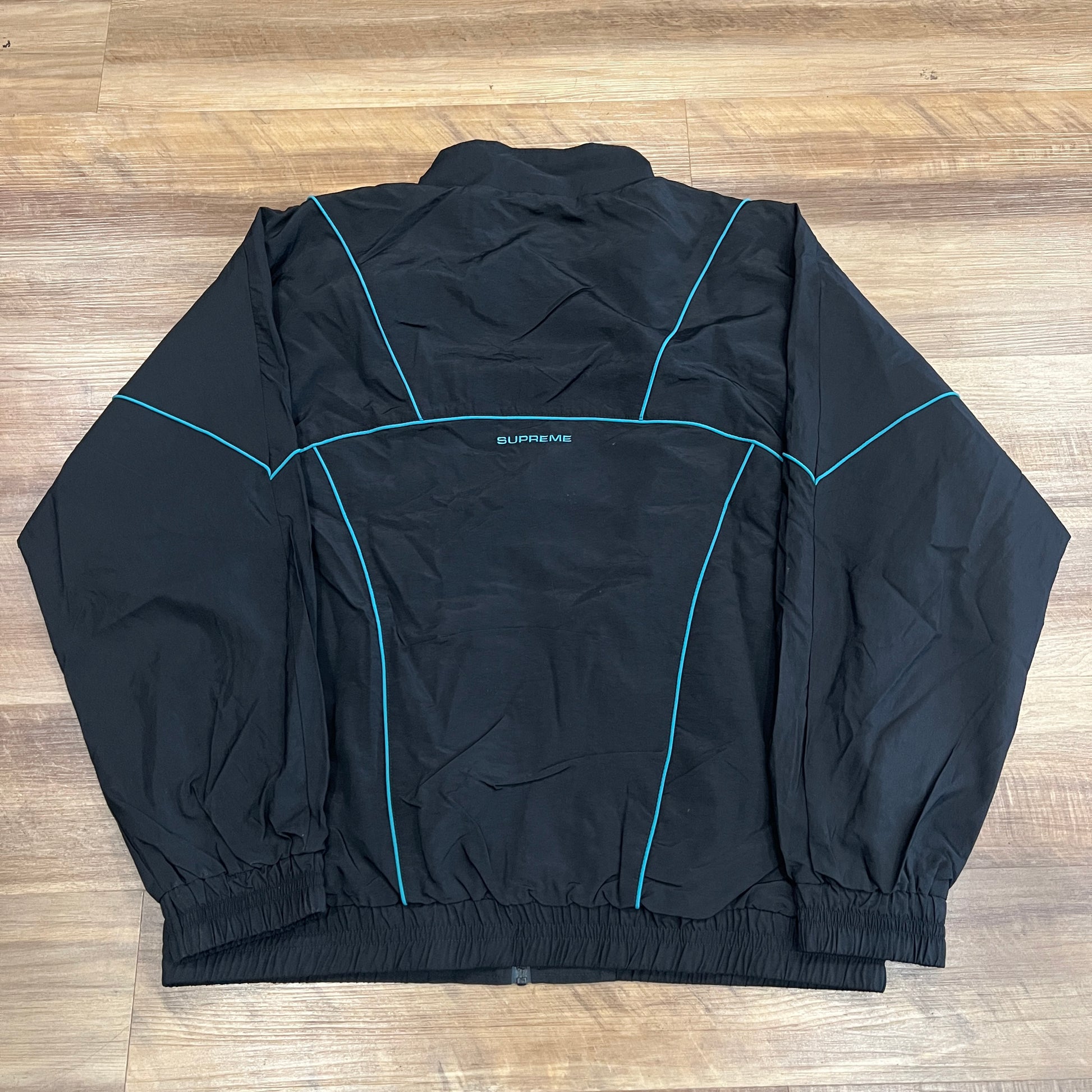 The Supreme Piping Track Jacket (SS19) in black features teal piping, elastic cuffs, a zip-up front, and a subtle logo on the left chest. The extra-large jacket is displayed laid flat on a wooden floor.