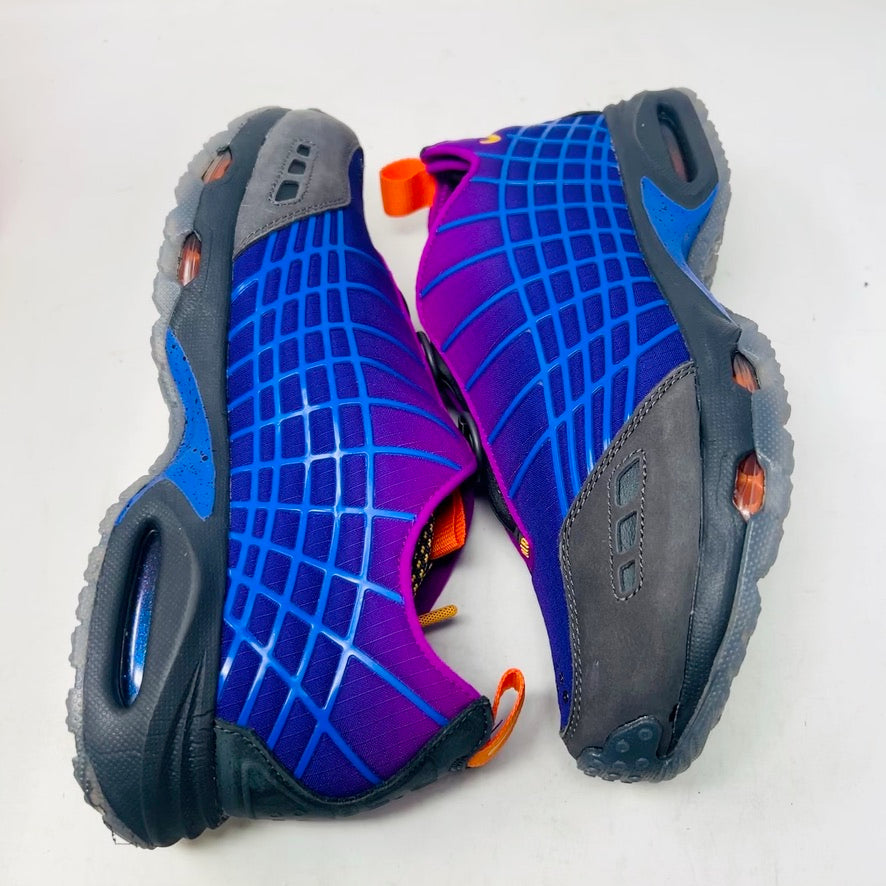 Displayed on an orange Nike shoe box is the colorful Nike Air Max Sunder SP Kids of Immigrants Sun Down sneaker, featuring a grid pattern with shades of purple, blue, and gray, complemented by orange accents and a swoosh logo. The shoe box shows a k-i logo inside a circle.