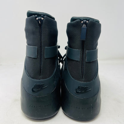 The Nike Air Fear of God 1 Triple Black is a high-top, dark, minimalist sneaker with angular overlays and a textured sole. It features a high collar, matte finish, dark laces, and has a small logo near the midsole. The background is light gray with general wear signs.