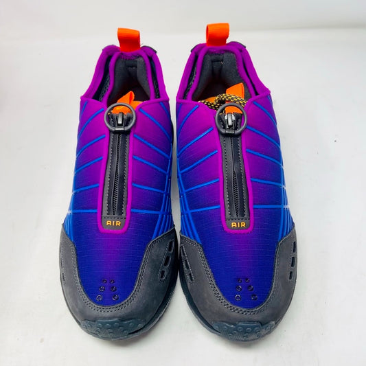 Displayed on an orange Nike shoe box is the colorful Nike Air Max Sunder SP Kids of Immigrants Sun Down sneaker, featuring a grid pattern with shades of purple, blue, and gray, complemented by orange accents and a swoosh logo. The shoe box shows a k-i logo inside a circle.