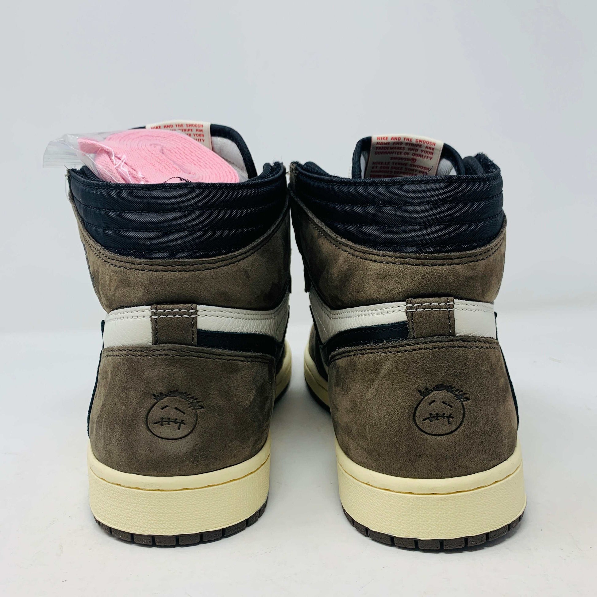A pair of authentic Jordan 1 High Travis Scott Mocha sneakers in brown, white, and black is displayed on an orange and brown Nike shoebox. These feature a distinctive reversed black Swoosh and come with extra laces for versatile styling.