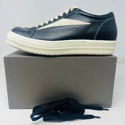 The Rick Owens Vintage Sneaks Black Milk from Holy Ground Sneaker Shop are showcased on a sleek gray box. The black leather sneakers with white accents have a thick white sole and come with black laces in front, offering style straight from the dust bag.