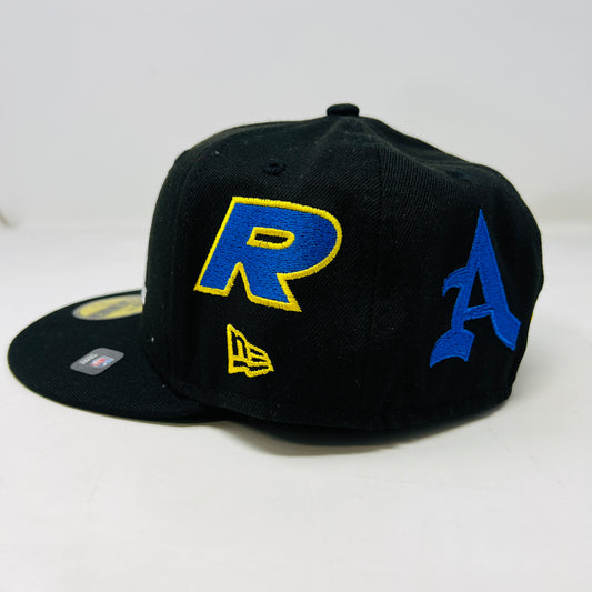 The New Era x Born x Raised x LA RAMS baseball cap by born x raised features LA in yellow and white embroidery on the front. It has a size 7 1/2 sticker on the brim with yellow and blue side patches, all set against a plain white background for a bold statement.
