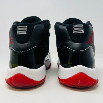 A pristine black and red Jordan 11 Retro Playoffs Bred (2019) (GS) sneaker is on an orange Nike box, featuring a patent leather upper, white midsole, red outsole, and the Jumpman logo near the heel. The label indicates year and size details.