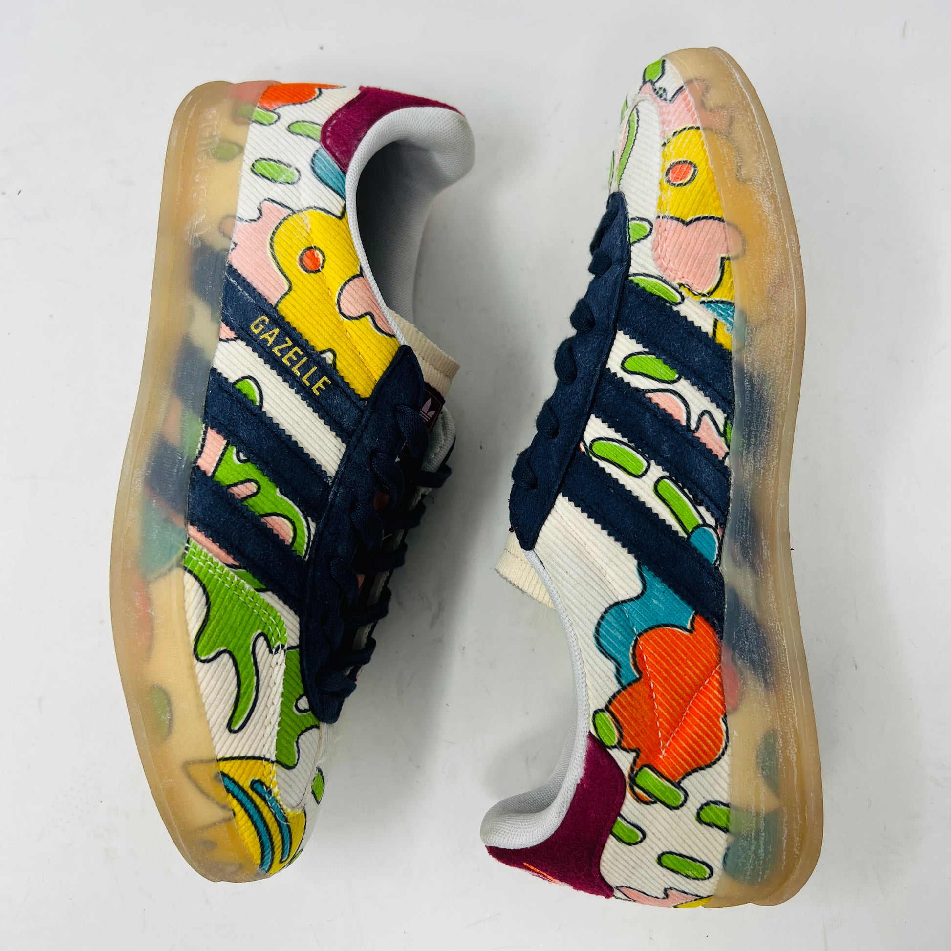The Adidas Gazelle Indoor Sean Wotherspoon sneaker, with colorful abstract uppers in yellow, red, pink, and green, dark blue stripes, red laces, and translucent soles, rests on a blue box showing size and product details.