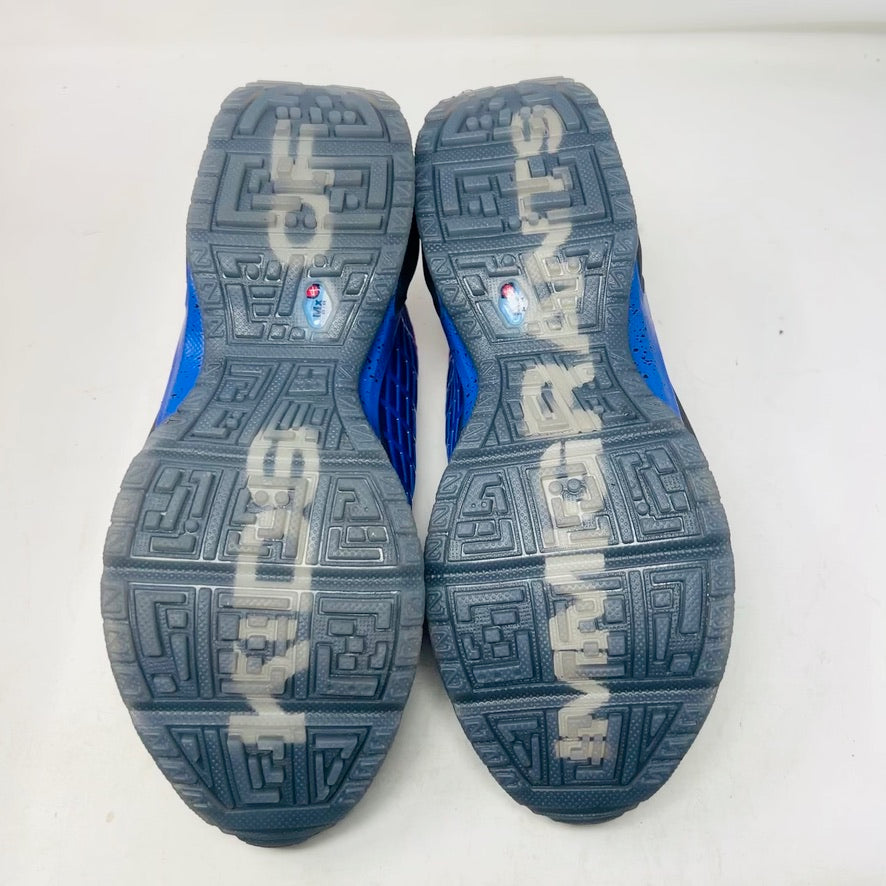 Displayed on an orange Nike shoe box is the colorful Nike Air Max Sunder SP Kids of Immigrants Sun Down sneaker, featuring a grid pattern with shades of purple, blue, and gray, complemented by orange accents and a swoosh logo. The shoe box shows a k-i logo inside a circle.