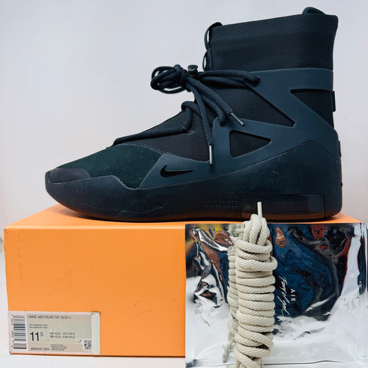 A black high-top Nike Air Fear of God 1 Triple Black sneaker, size 11.5, is on an orange box. The uppers show light wear, and OG accessories include white shoelaces and a metallic bag on the side.