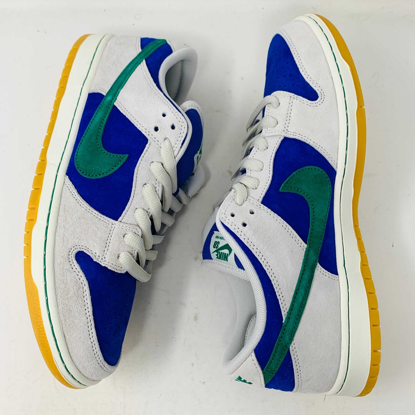The Nike SB Dunk Low Hyper Royal Malachite, a fresh pair of white and blue sneakers with a green swoosh and gum sole, includes extra laces and is presented atop a purple Nike SB shoe box.
