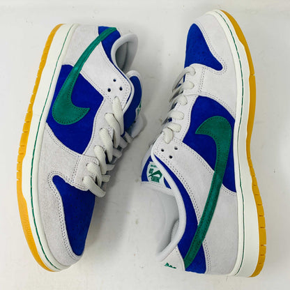 The Nike SB Dunk Low Hyper Royal Malachite, a fresh pair of white and blue sneakers with a green swoosh and gum sole, includes extra laces and is presented atop a purple Nike SB shoe box.