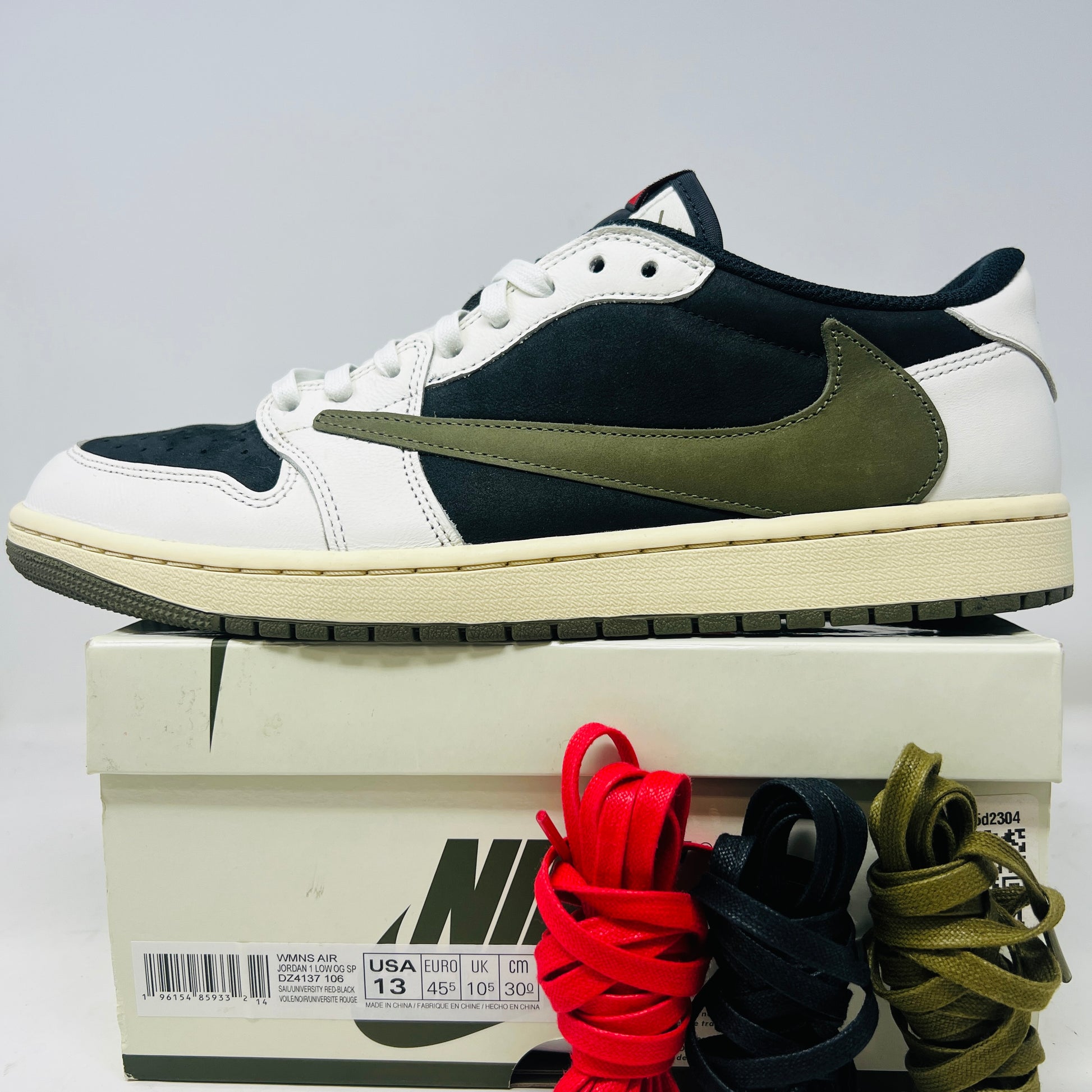 The Jordan 1 Retro Low OG SP Travis Scott Olive (Womens) sneakers, featuring clean uppers with a prominent Swoosh and hues of black, white, and olive green, rest on a Nike box. Included are red, black, and olive green laces. Light wear is evident with slight heel drag on each shoe.