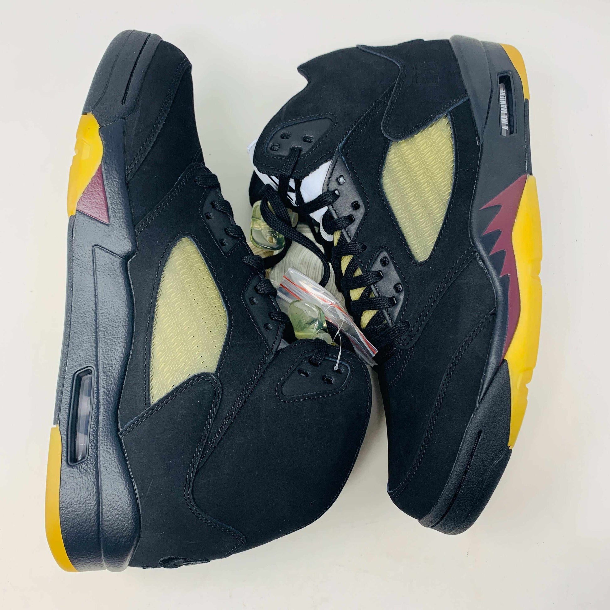The Jordan 5 Retro A Ma Maniére Dusk sneakers feature a black high-top design with yellow mesh side panels, and colorful yellow and maroon accents on the soles. Displayed on a white background, they have black laces complemented by a transparent lace holder.