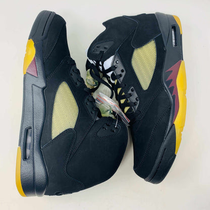 The Jordan 5 Retro A Ma Maniére Dusk sneakers feature a black high-top design with yellow mesh side panels, and colorful yellow and maroon accents on the soles. Displayed on a white background, they have black laces complemented by a transparent lace holder.