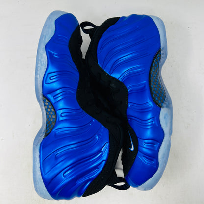 A brand-new Nike Air Foamposite One International Blue sneaker, featuring a striking blue upper with black accents, rests on a sleek black Nike shoebox. It has black mesh laces, a translucent sole with carbon fiber plate, and an iconic side swoosh.