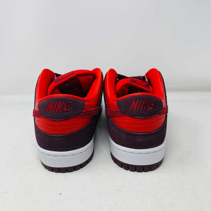 A 100% authentic, brand new Nike SB Dunk Low Cherry sneaker in red and dark brown sits on a branded purple Nike SB shoebox. It features a red swoosh, toe box, dark brown overlays, extra laces, white midsole, and dark brown outsole.