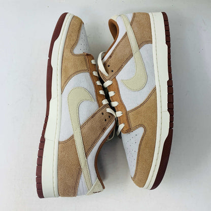 A brand new pair of Nike Dunk Low Medium Curry sneakers, with a beige suede upper and white swoosh accents, rests in pristine condition on a red box featuring the bold white Nike logo, set against a plain backdrop.