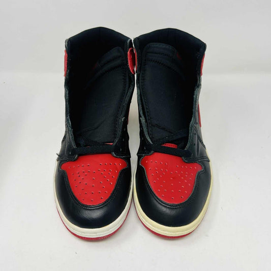 A pair of brand new Jordan 1 Retro Bred (1994) sneakers is displayed on a box, featuring the iconic Nike swoosh and wings logo. A black-and-white photo of the shoes is behind them. The box has various sneaker-related graphics, emphasizing their authenticity.