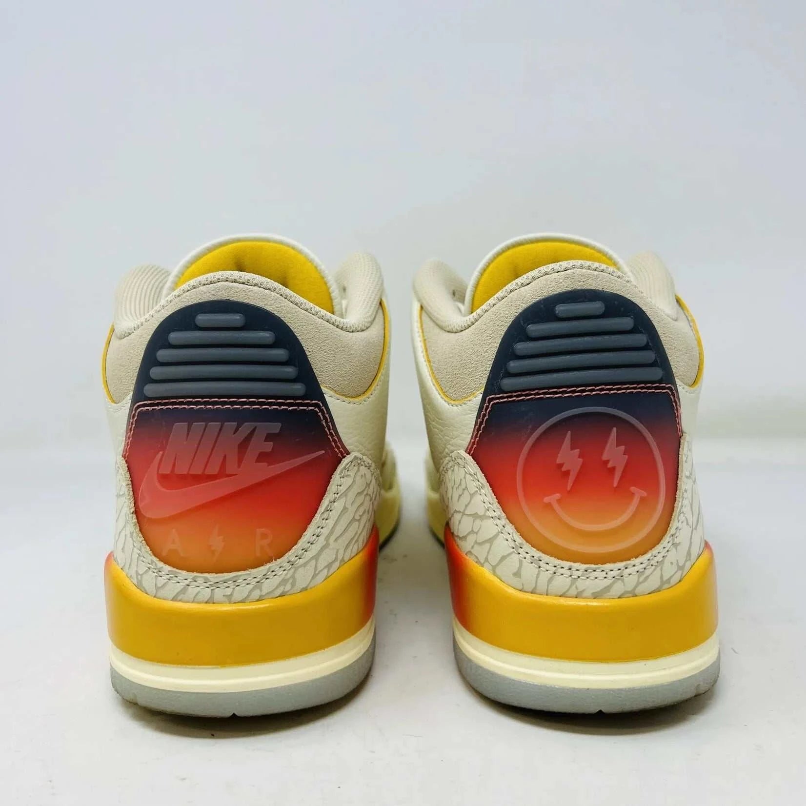The Jordan 3 Retro SP J Balvin Medellín Sunset showcases clean cream uppers atop a shoebox with a gradient from yellow to dark red. A smiley face tag with lightning bolts confirms its 100% authenticity, highlighting this special 2023 release.