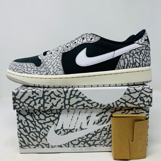 The Jordan 1 Retro Low OG Black Cement sneaker, with black and gray elephant print and a white swoosh, is showcased on a matching shoe box. A small brown box with extra laces is beside it, all set against a plain background.