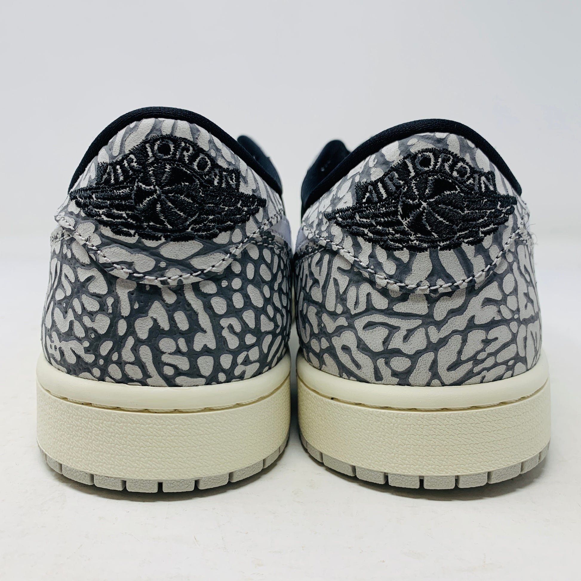 The Jordan 1 Retro Low OG Black Cement sneaker, with black and gray elephant print and a white swoosh, is showcased on a matching shoe box. A small brown box with extra laces is beside it, all set against a plain background.