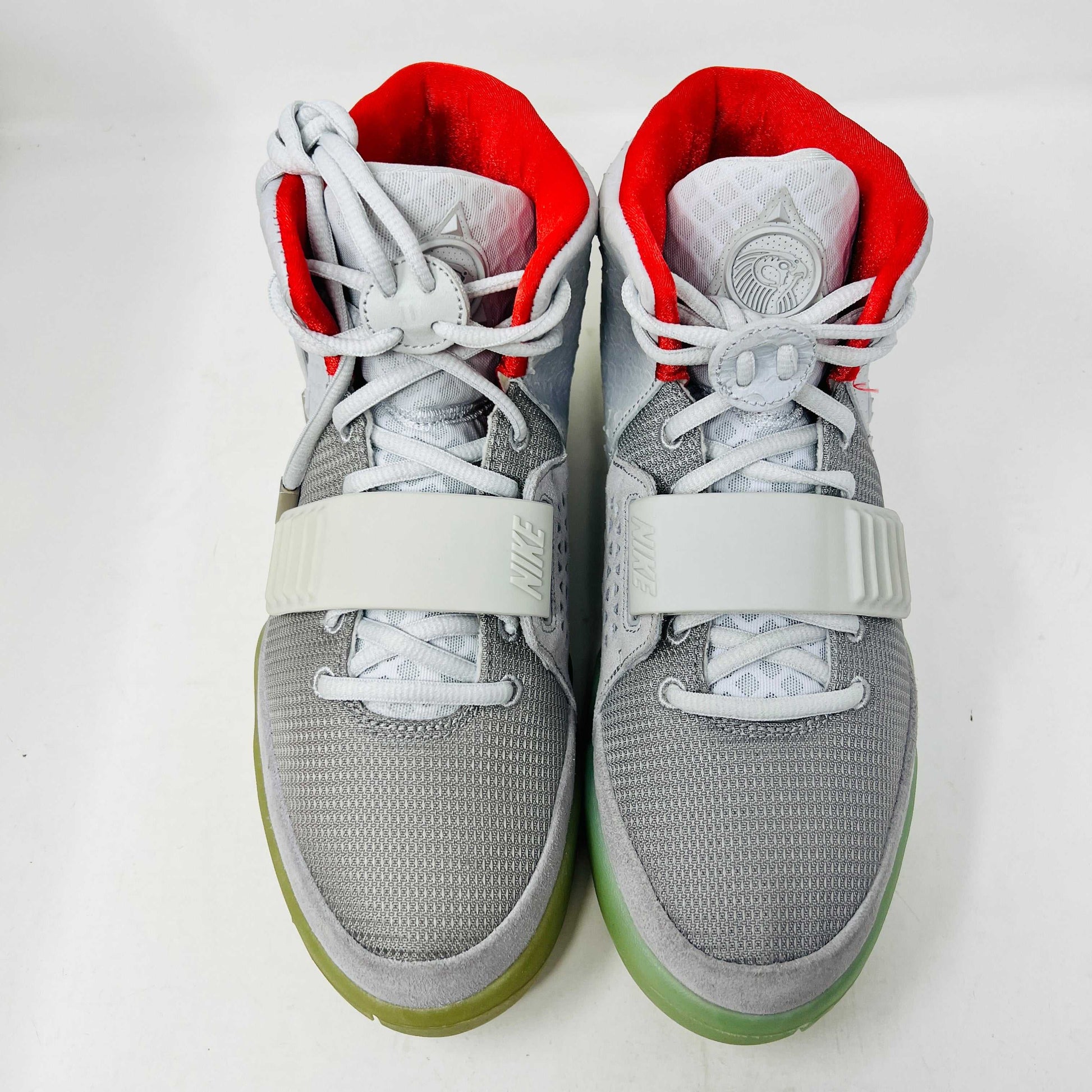 A pair of new Nike Air Yeezy 2 Pure Platinum high-tops rests on a cardboard box. The sneakers are mostly gray with a red lining, midfoot strap, and green-tinted soles. The label reads Air Yeezy 2 NRG, size 9, showcasing authentic detailing by Nike.