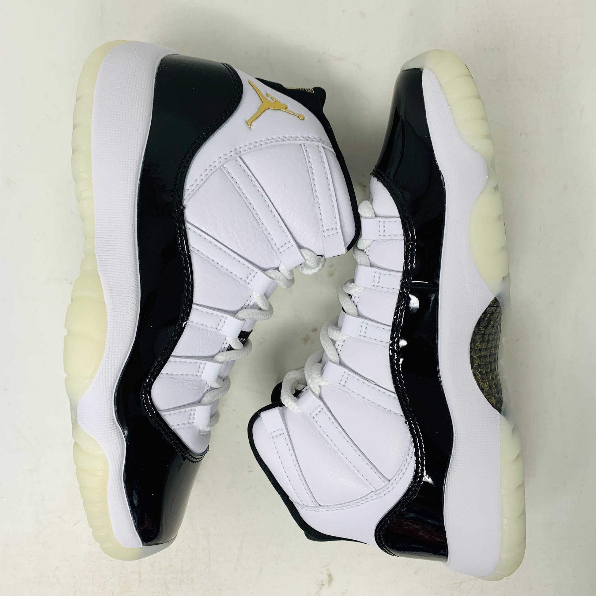 A pair of authentic Jordan 11 Retro DMP Gratitude (2023) (GS) sneakers, featuring a glossy white and black finish with a gold logo, is placed on top of a gray Nike Air shoebox.