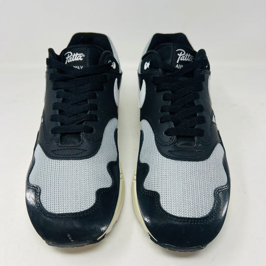 The Nike Air Max 1 Patta Waves Black sneakers are presented on their box, showcasing clean uppers with gray and white accents. They feature a bracelet-like chain tag with a flag detail, and white laces gracefully draped in front.