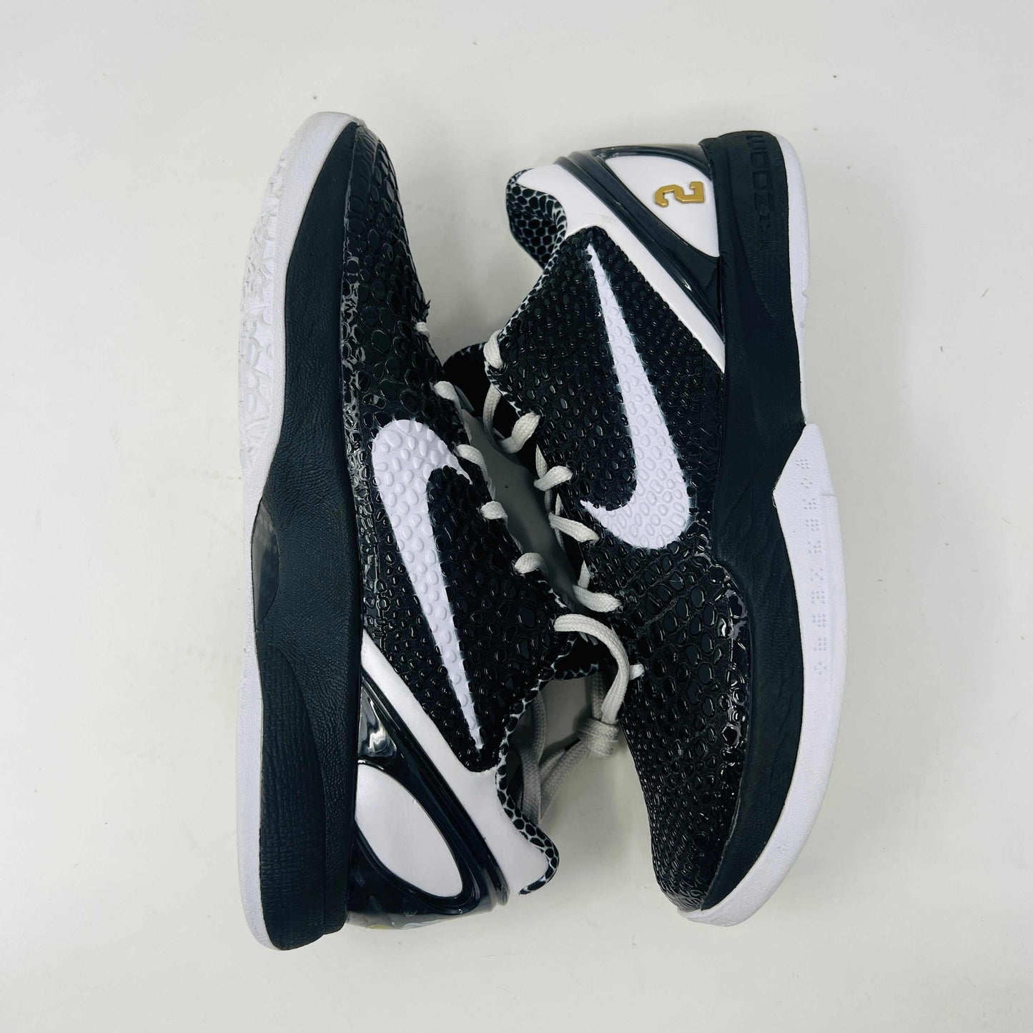 The black and white Nike Kobe 6 Protro Mambacita Sweet 16 shoe features a glossy finish, a gold number 2 on the heel, and light wear with a minor scratch on the back tab. It comes in sizes 8 and 9.5, displayed on a black box labeled Kobe V Proto, and is guaranteed authentic by KOBE.