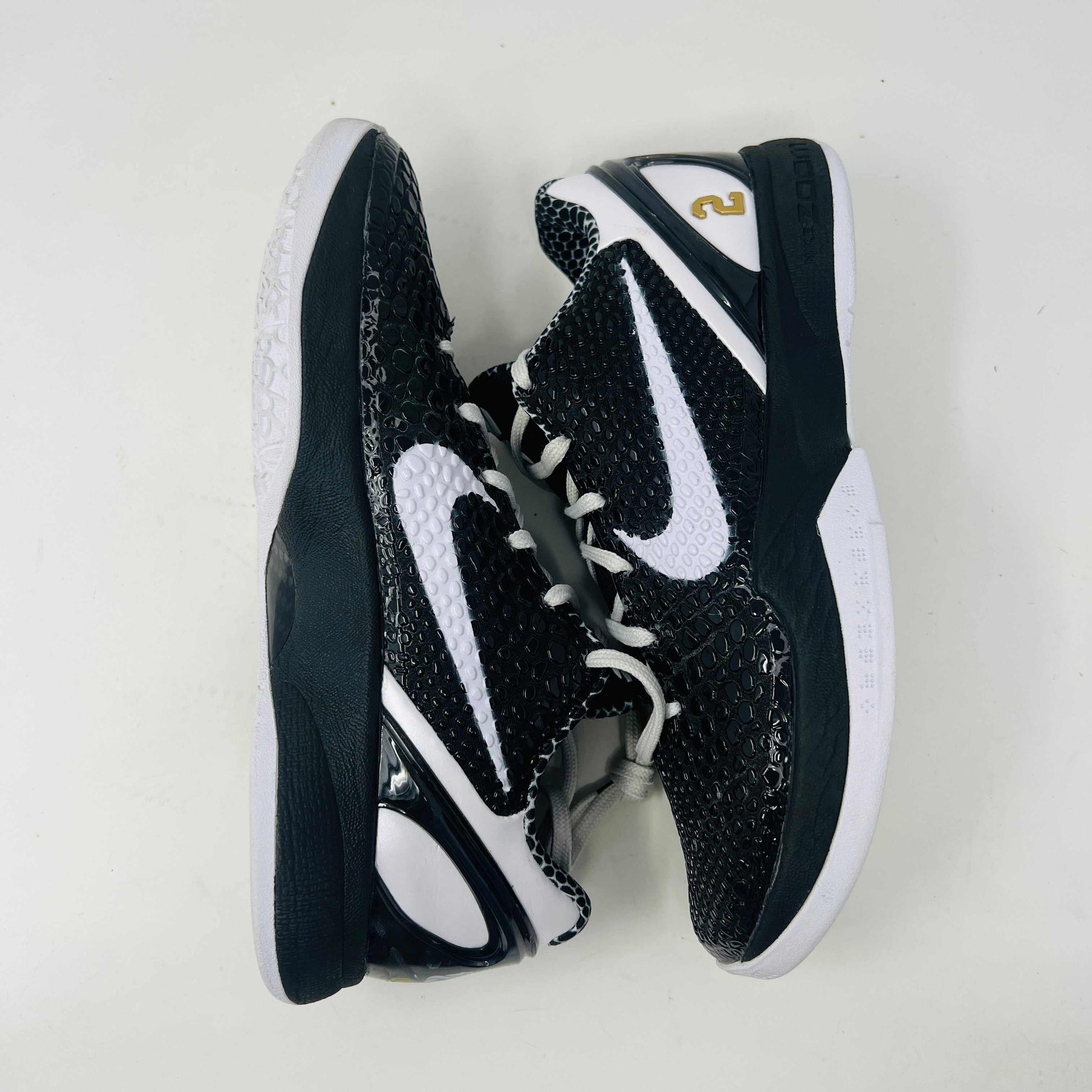 The black and white Nike Kobe 6 Protro Mambacita Sweet 16 shoe features a glossy finish, a gold number 2 on the heel, and light wear with a minor scratch on the back tab. It comes in sizes 8 and 9.5, displayed on a black box labeled Kobe V Proto, and is guaranteed authentic by KOBE.
