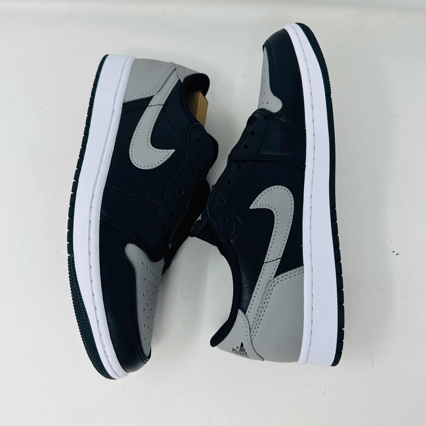 A pair of Jordan 1 Low OG Shadow (2024) sneakers, in black and gray, rests atop a gray shoe box with a large black Nike logo. Beside the box is a small brown cardboard package featuring a cutout revealing shoelaces inside.