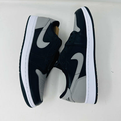 A pair of Jordan 1 Low OG Shadow (2024) sneakers, in black and gray, rests atop a gray shoe box with a large black Nike logo. Beside the box is a small brown cardboard package featuring a cutout revealing shoelaces inside.