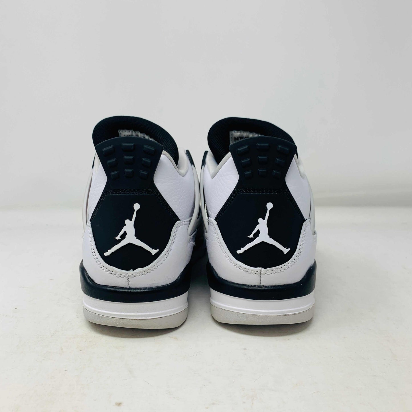 The Jordan 4 Military Black GS sneakers, featuring distinctive side netting and a logo on the tongue, are brand new and authentic in white and gray with black accents. They face forward on a white surface against a plain background, embodying modern style.