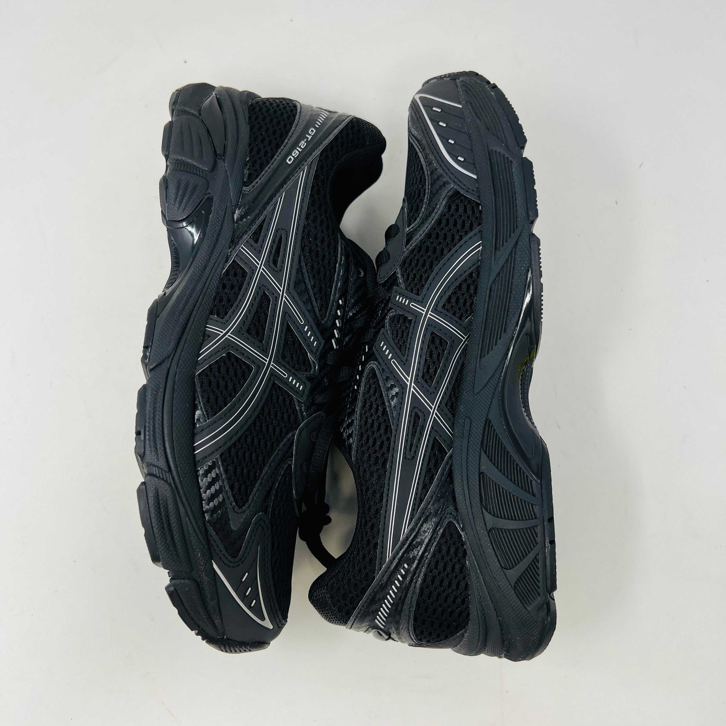 The new ASICS GT-2160 JJJJound Black running shoe sits on its box, featuring extra laces in a clear bag. These authentic shoes present a sleek design with gray detailing and a side text label.