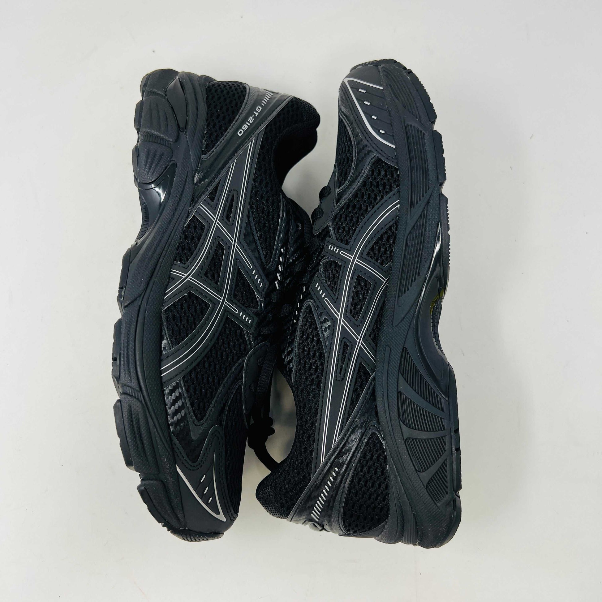 The new ASICS GT-2160 JJJJound Black running shoe sits on its box, featuring extra laces in a clear bag. These authentic shoes present a sleek design with gray detailing and a side text label.