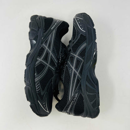 The new ASICS GT-2160 JJJJound Black running shoe sits on its box, featuring extra laces in a clear bag. These authentic shoes present a sleek design with gray detailing and a side text label.
