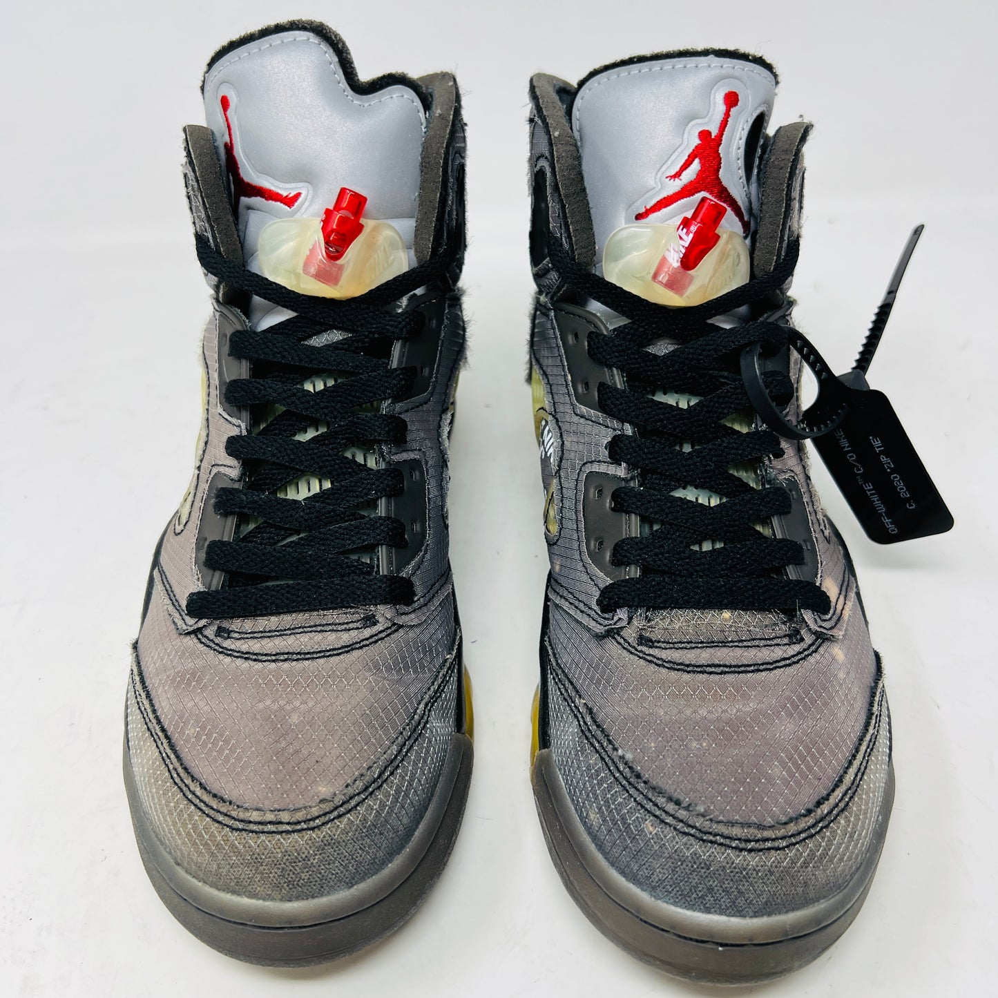 The Jordan 5 Retro Off-White Muslin high-top sneaker, featuring a transparent mesh window and 23 on the heel, is displayed on its box with a white and red shoelace packet in front. A small bleach stain adds unique character to the design.