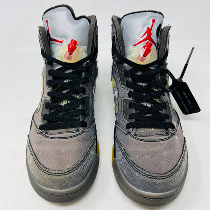 The Jordan 5 Retro Off-White Muslin high-top sneaker, featuring a transparent mesh window and 23 on the heel, is displayed on its box with a white and red shoelace packet in front. A small bleach stain adds unique character to the design.