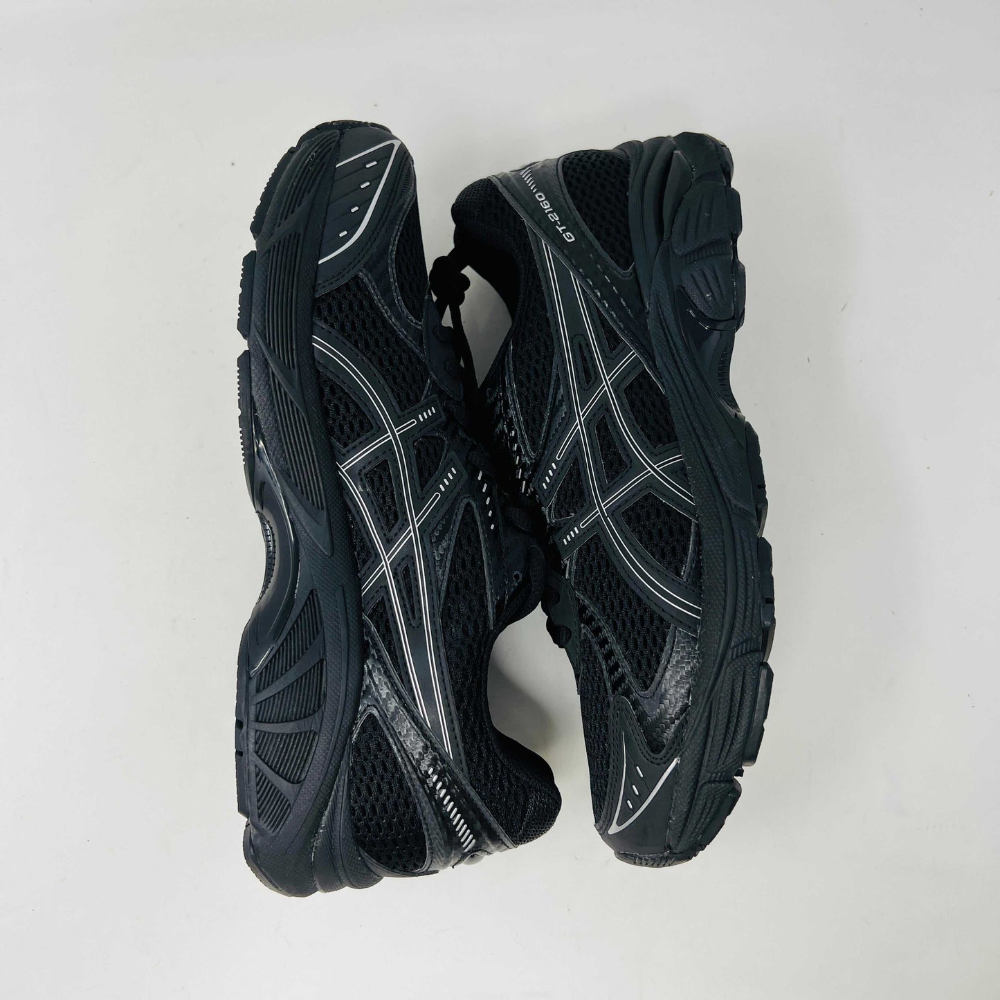 The new ASICS GT-2160 JJJJound Black running shoe sits on its box, featuring extra laces in a clear bag. These authentic shoes present a sleek design with gray detailing and a side text label.
