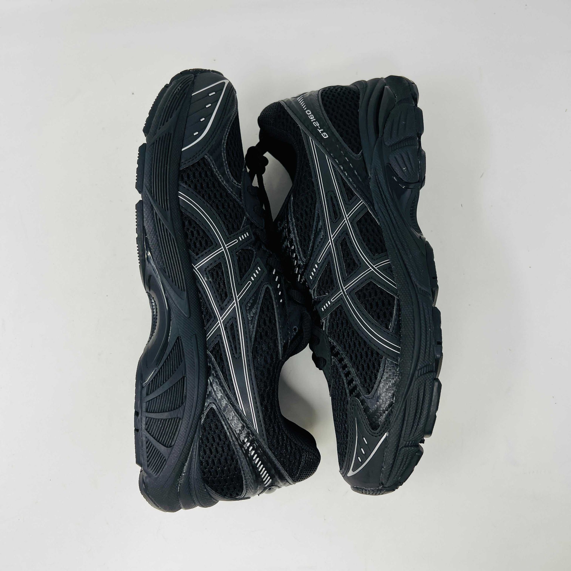 The new ASICS GT-2160 JJJJound Black running shoe sits on its box, featuring extra laces in a clear bag. These authentic shoes present a sleek design with gray detailing and a side text label.