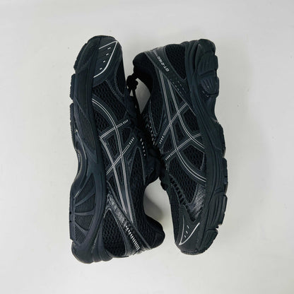 The new ASICS GT-2160 JJJJound Black running shoe sits on its box, featuring extra laces in a clear bag. These authentic shoes present a sleek design with gray detailing and a side text label.