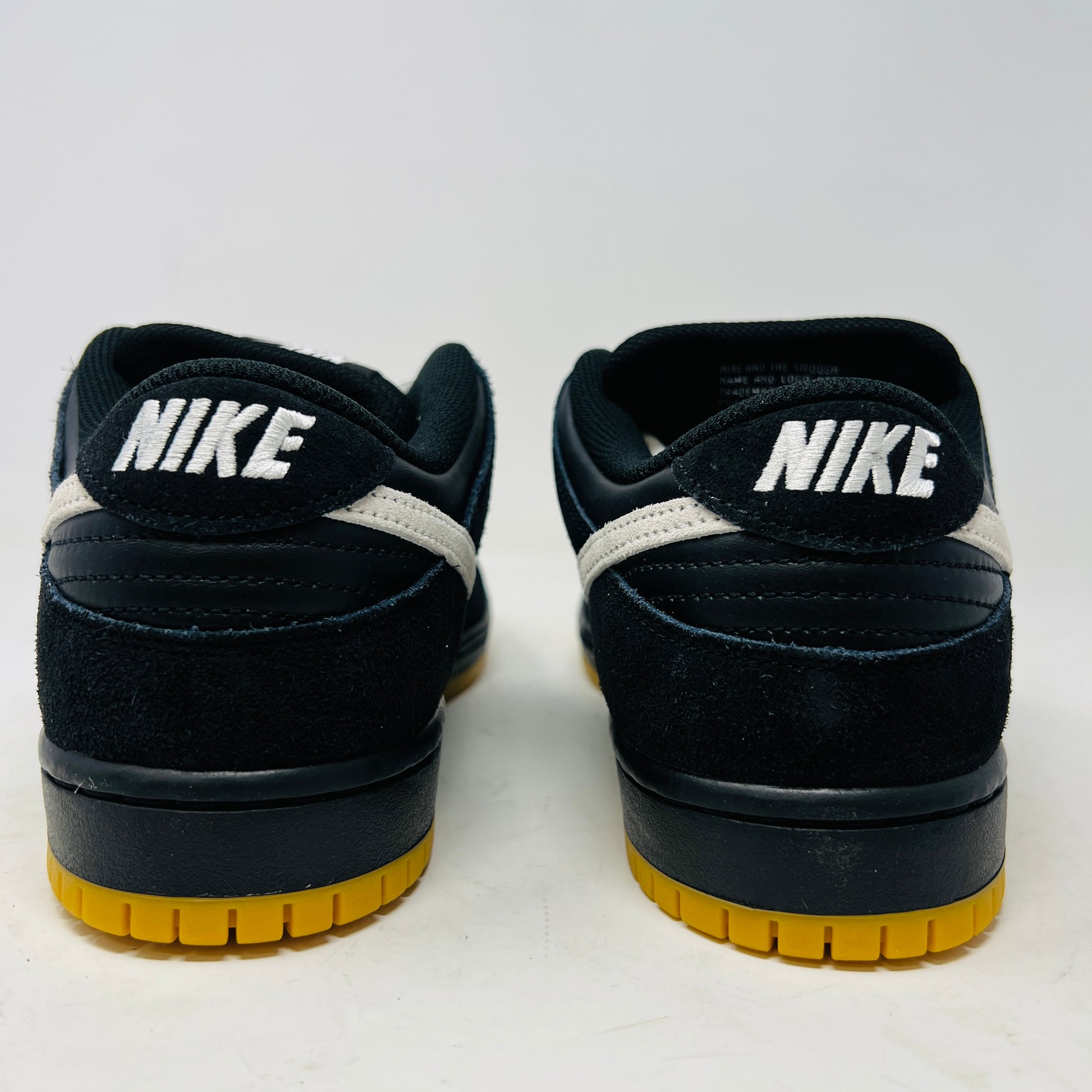 A black Nike Dunk Low Pro SB Black White Gum sneaker with white laces and swoosh sits on a purple Nike SB box. The 2025 shoe has a yellow sole, leather-texture finish, and both are in new condition.