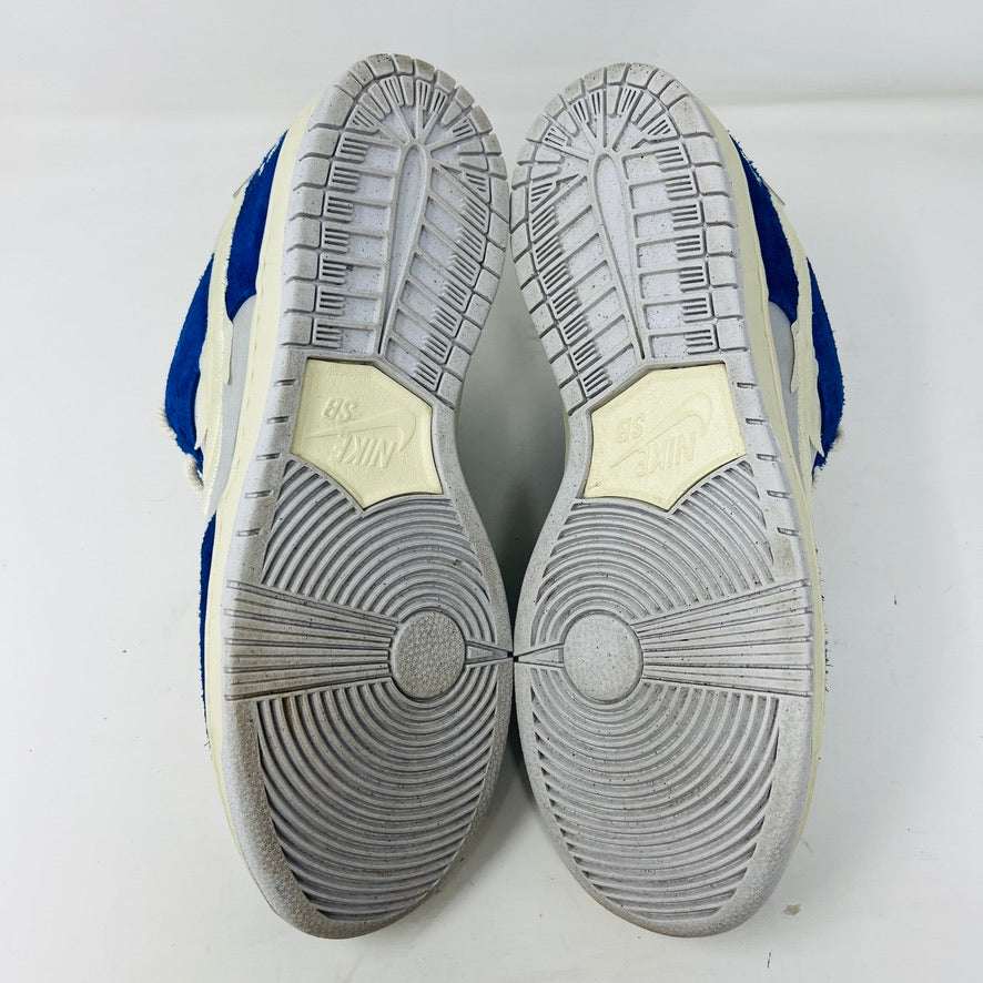 The soles of a pre-owned pair of Nike SB Dunk Low Pro Fly Streetwear Gardenia sneakers face up, featuring circular grip patterns and the iconic Nike logo. Primarily blue and white, these are sold as-is with no returns; all sales are final.