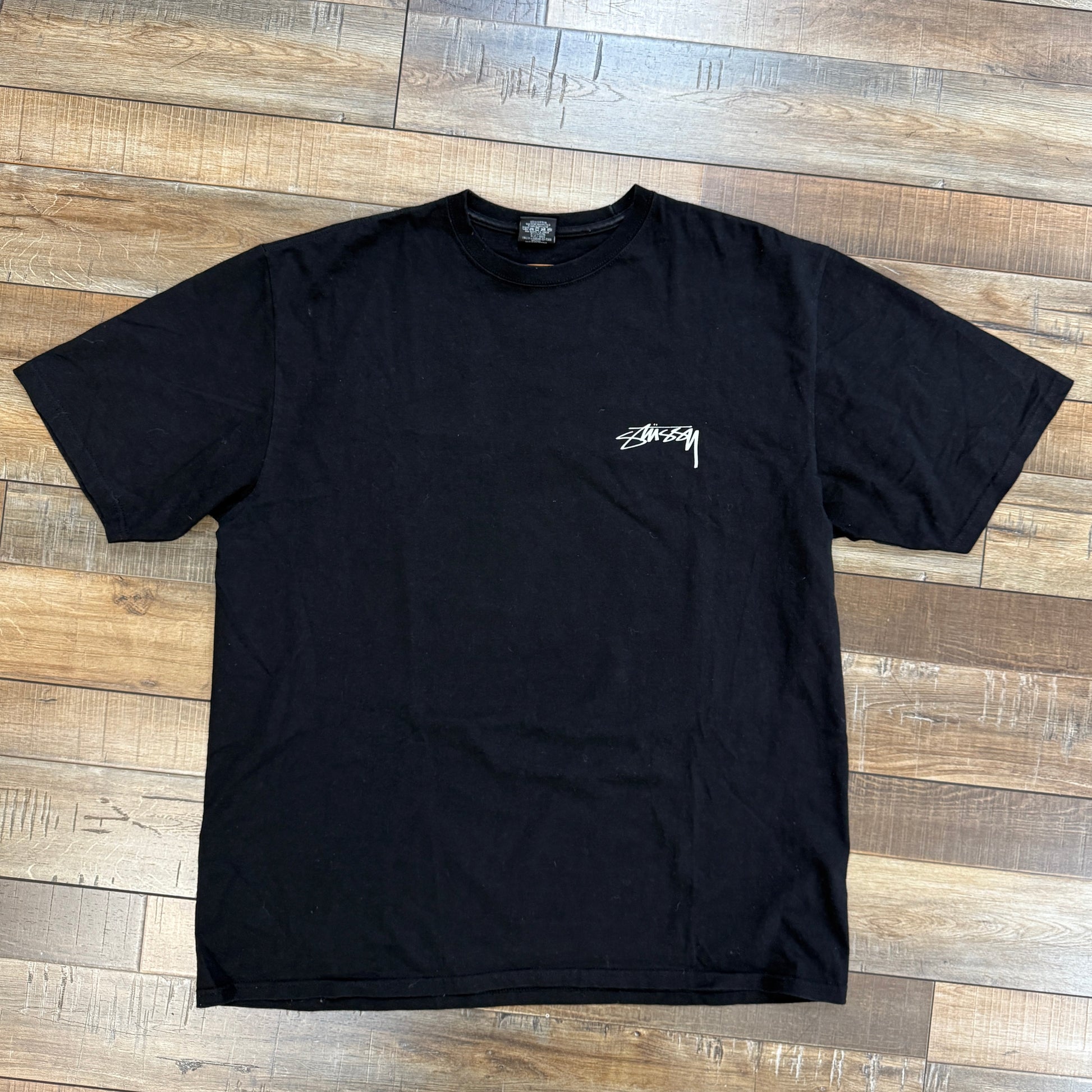 Preowned black Stussy Sphinx Tee with logo on chest laid flat on wooden floor