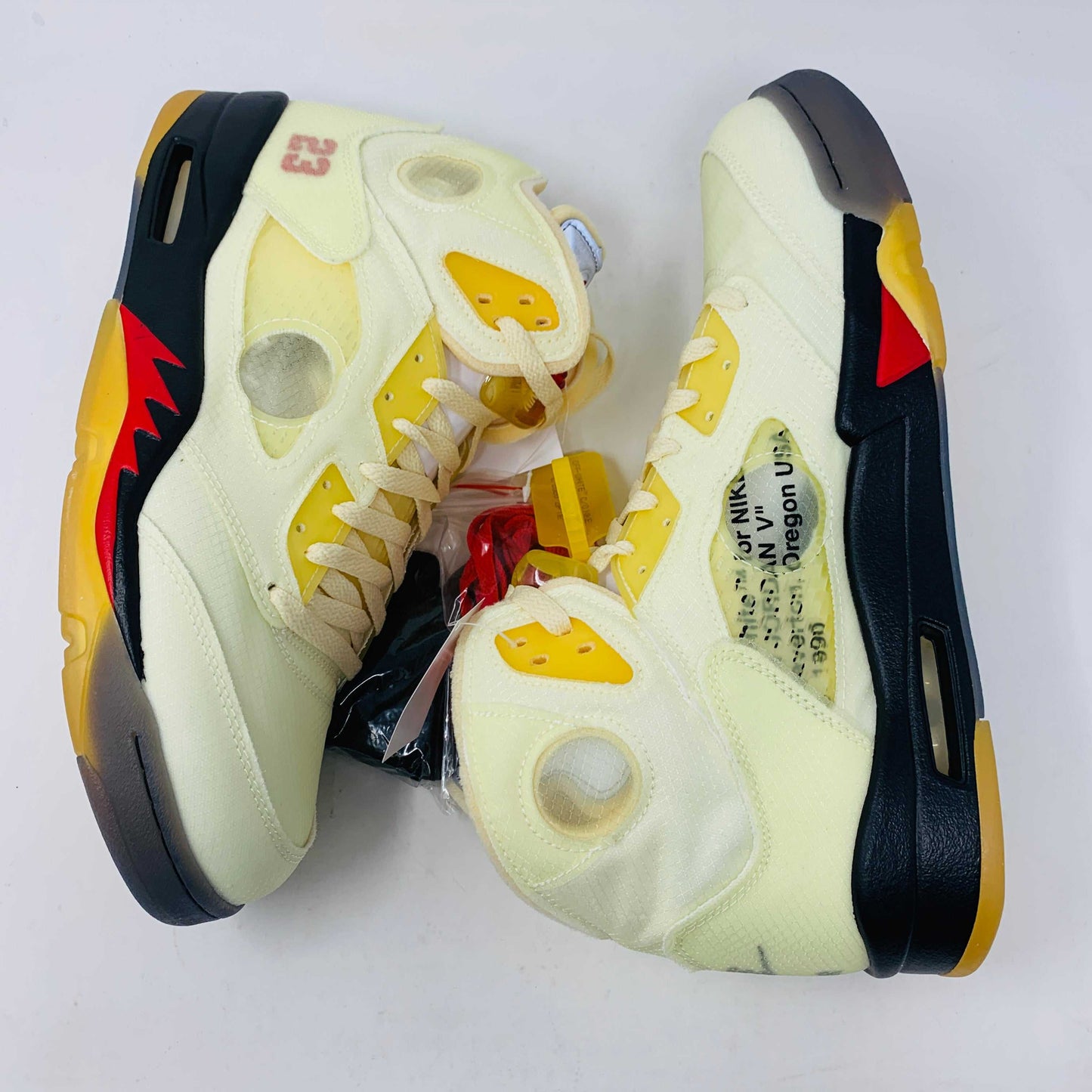 The Jordan 5 Off White Sail sneakers feature a yellow and black design with red accents, a visible air unit, and the number 23 on the side. BRAND NEW in a black shoebox with a red Jumpman logo and speckled base, they include an attached yellow tag and come with EXTRA LACES for versatility.