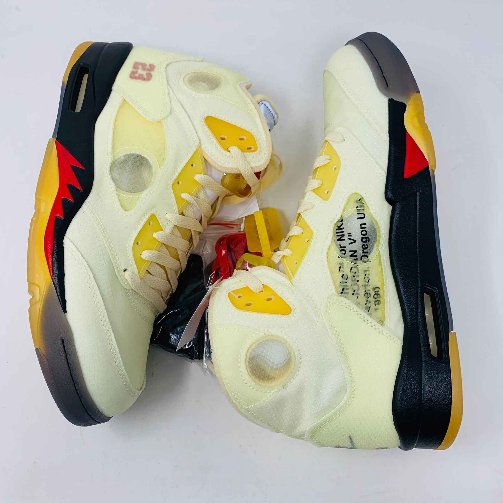 The Jordan 5 Off White Sail sneakers feature a yellow and black design with red accents, a visible air unit, and the number 23 on the side. BRAND NEW in a black shoebox with a red Jumpman logo and speckled base, they include an attached yellow tag and come with EXTRA LACES for versatility.